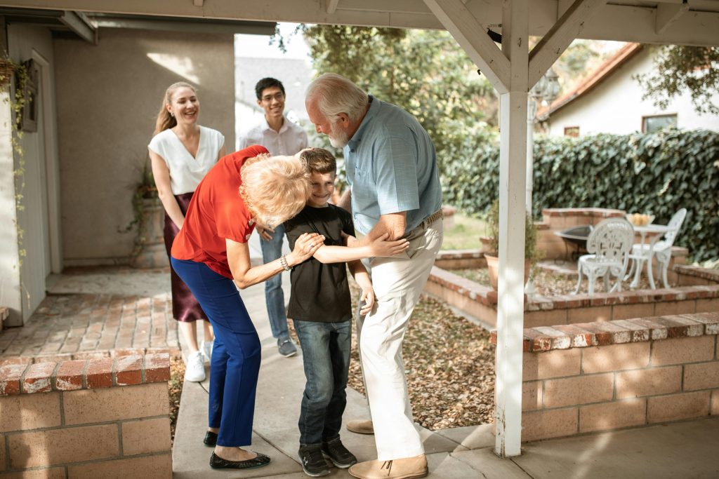 eine Familie mit drei Generationen im Garten, die Großeltern umarmen ihren Enkel, die Eltern gehen von hinten auf die drei zu